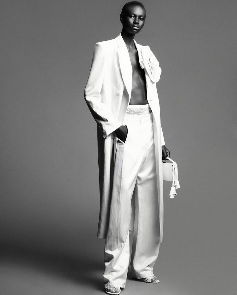 Elegant studio shot of Black female fashion model in white oversized pantsuit, floral shoulder detail, box purse, and gray backdrop.