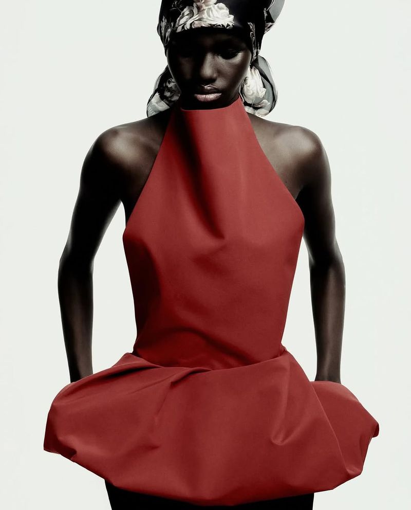 Dark-skinned adult woman in a red halter top and patterned headscarf, posing in front of a plain white studio backdrop with hands at her hips.