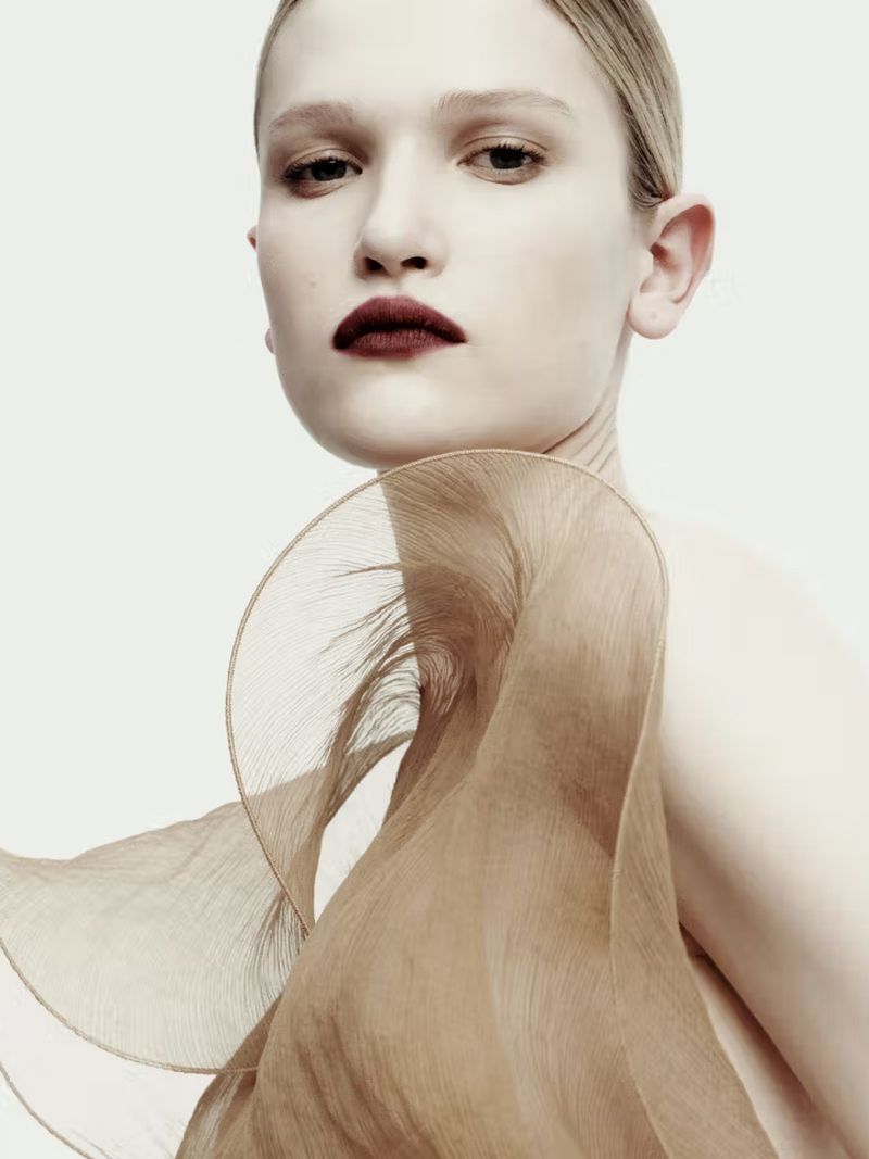 Close-up portrait of a female fashion model with dark lipstick and a tan fabric accessory on a white background in a photo studio.