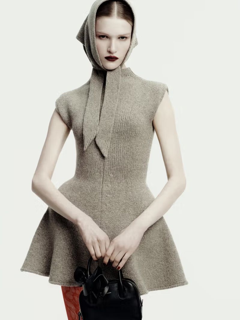 Model wearing a grey knitted peplum dress with a matching headscarf and holding a black handbag against a plain background