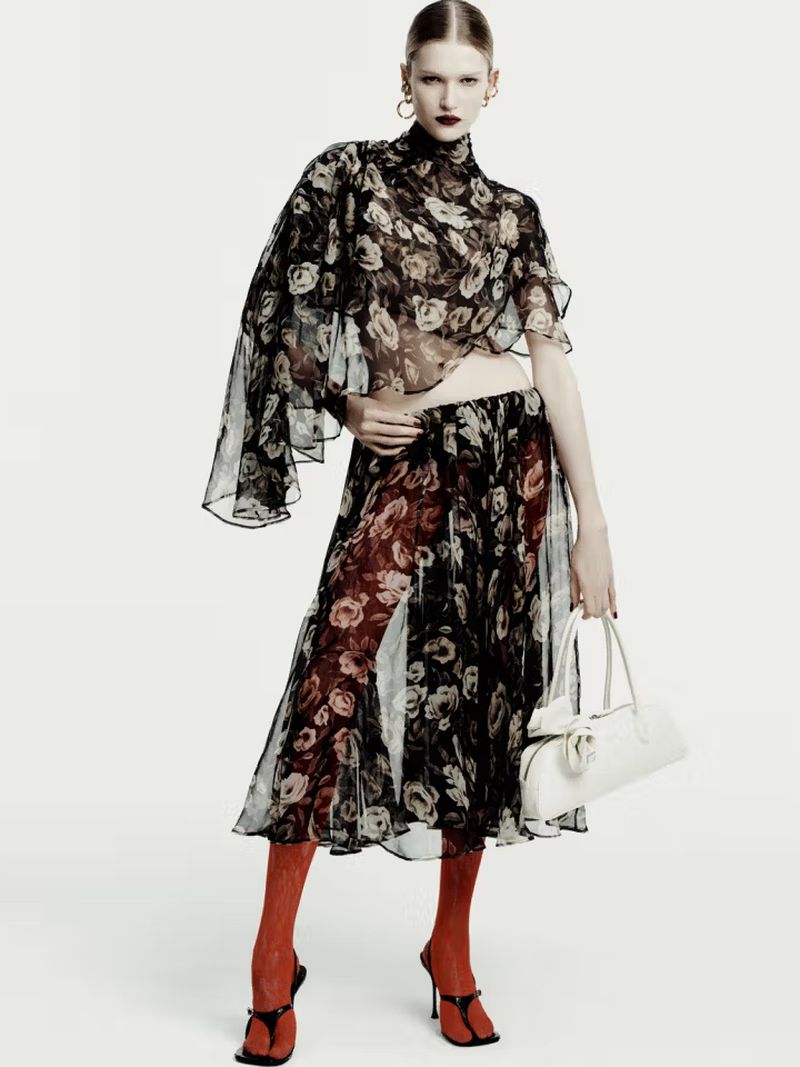 A fair-skinned fashion model in a black floral top and skirt, red stockings, and black heels stands in a photo studio with a white bag.