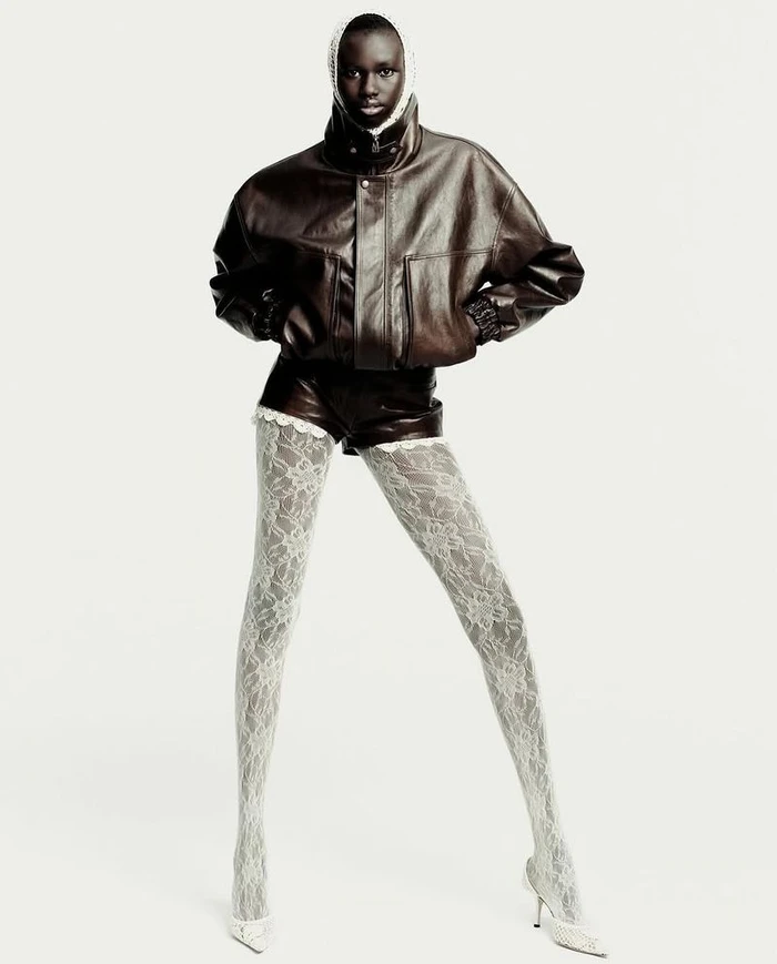 Model wearing a dark brown leather jacket and shorts with white lace tights against a plain white background.