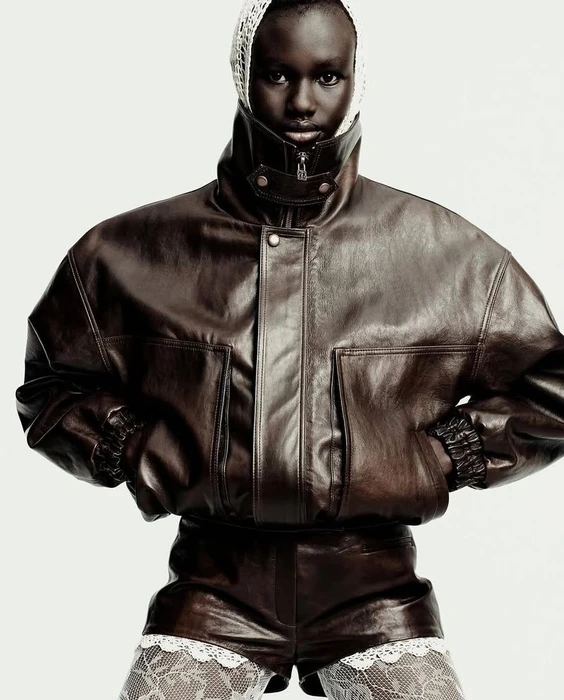 Model in brown leather jacket and matching shorts wearing a white lace head covering against a plain white background.