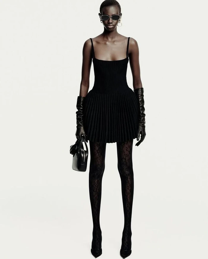 Model in black dress with pleated skirt, leather gloves, and sunglasses standing against a plain white background