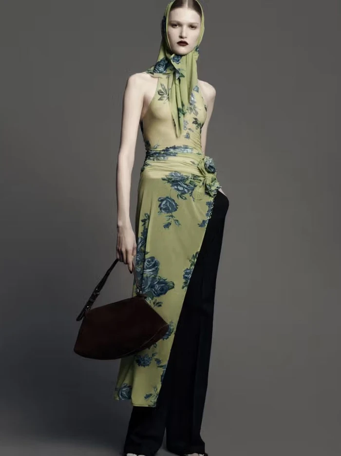 Model in green floral halter dress with matching headscarf and dark trousers holding a brown handbag against a grey seamless backdrop