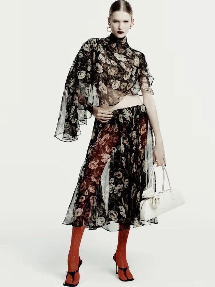 Model in a floral sheer top and matching skirt holding a white handbag against a seamless white background.