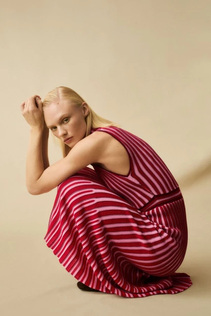 Woman in a pink and red striped dress squats in a neutral setting, showcasing fashion and style with a studio background.