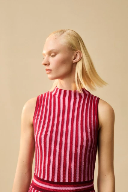Woman with blond hair models a pink and red striped sleeveless top and skirt against a neutral backdrop in a studio setting.