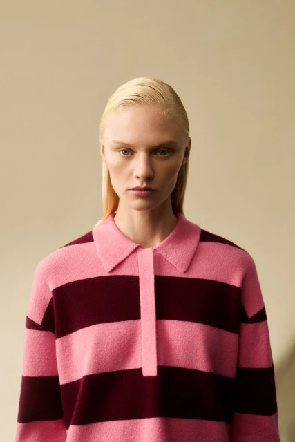 Fashion portrait of a blond woman in a pink and maroon striped, collared sweater against a neutral background in a studio setting.