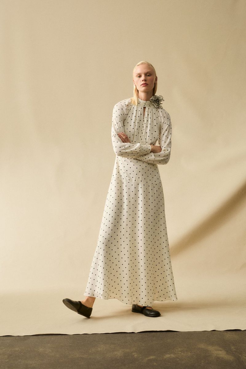 A slender, fair-skinned fashion model poses in a long-sleeved, polka-dot maxi dress, standing in a studio with arms crossed and legs slightly apart.