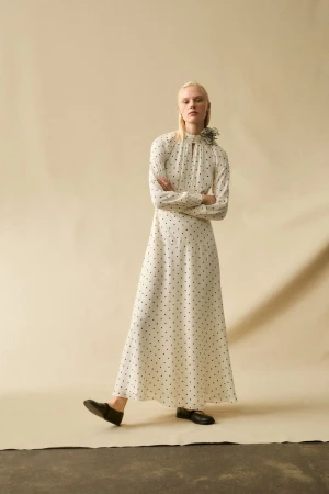 A slender, fair-skinned fashion model poses in a long-sleeved, polka-dot maxi dress, standing in a studio with arms crossed and legs slightly apart.