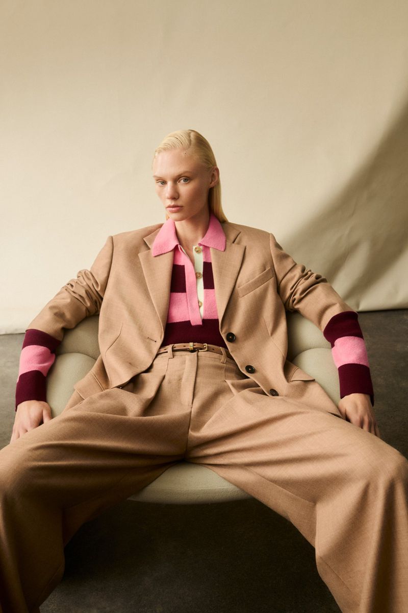 A fair-skinned woman with blonde hair sits on a green chair wearing a tan blazer, polo shirt, and slacks in a neutral studio.