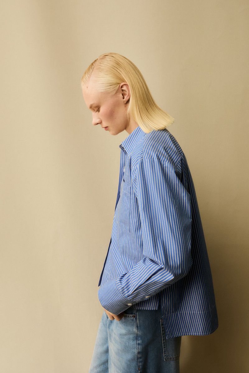 Blonde woman in blue and white striped button-down shirt and jeans in a studio setting, side profile with head tilted down.