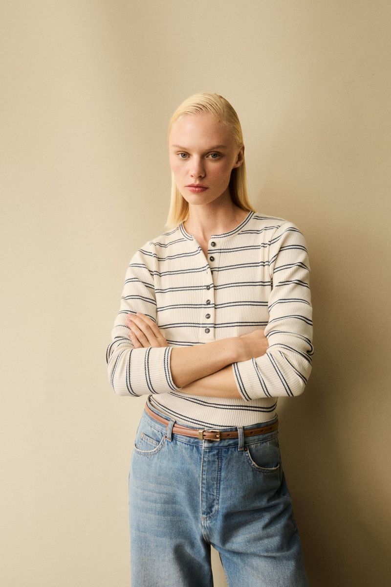 Blond woman in a striped long-sleeve shirt and wide-leg jeans poses with crossed arms against a neutral studio backdrop.