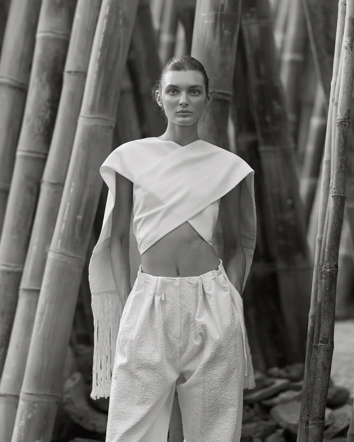 Black and white fashion shot of a woman in a light-colored top and pants posing in front of bamboo stalks outdoors, eyes looking forward.