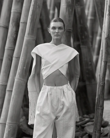 Black and white fashion shot of a woman in a light-colored top and pants posing in front of bamboo stalks outdoors, eyes looking forward.