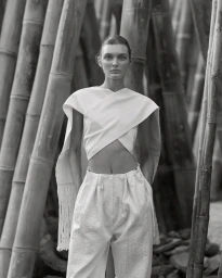 Model standing among tall bamboo stalks wearing a white draped crop top and high-waisted textured trousers in monochrome.