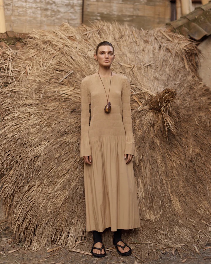 A woman in a tan dress stands against a haystack background, wearing sandals and a necklace, in an outdoor fashion shot.