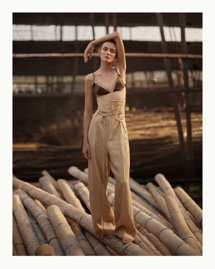 Fashion shot of a slender adult female posing in a brown bra top and beige high-waisted pants, standing on stacked bamboo poles outdoors.