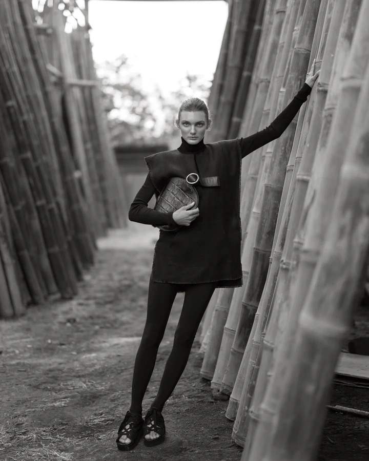Monochrome fashion portrait of a woman in dark dress and leggings holding a purse, standing amidst bamboo stalks in an outdoor setting.