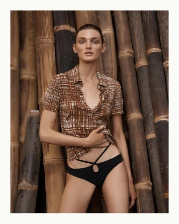 Fashion shot of a female model wearing a brown patterned top and black underwear posing in front of a bamboo background.