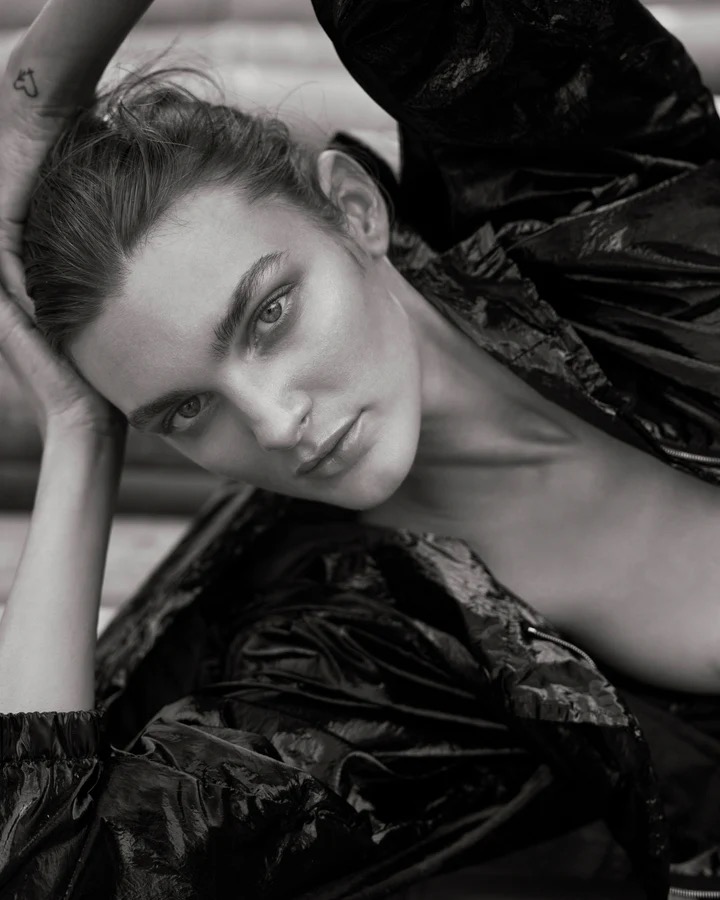 Striking black-and-white portrait of a young woman with distinctive features wearing a dark, shiny jacket, gazing directly at the camera.