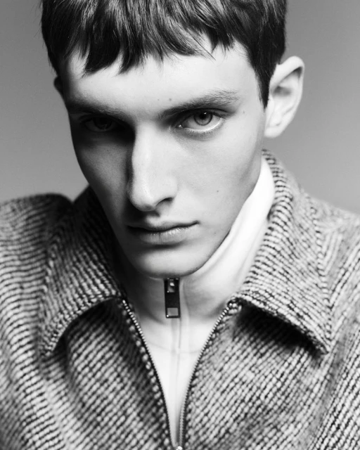 Monochrome portrait of a young adult man with short dark hair wearing a textured jacket over a zip-up top, against a plain background.