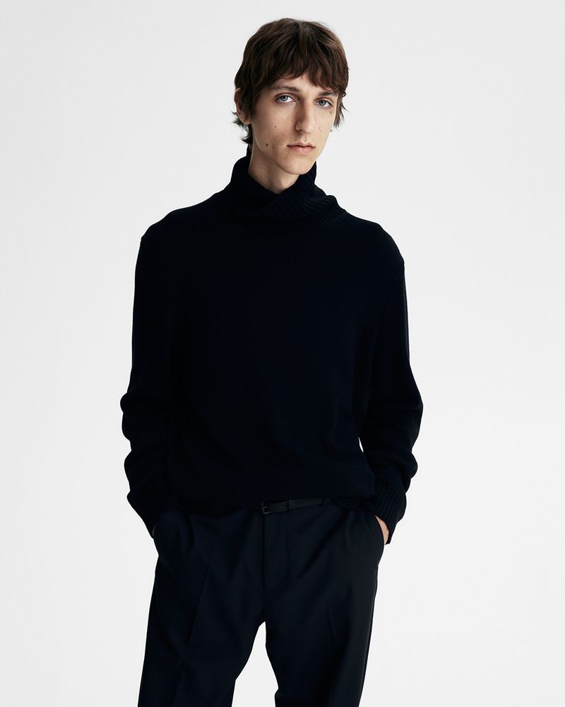 Studio portrait of a slender adult man in a dark turtleneck sweater, black pants, and a thin black belt posing on a white background.