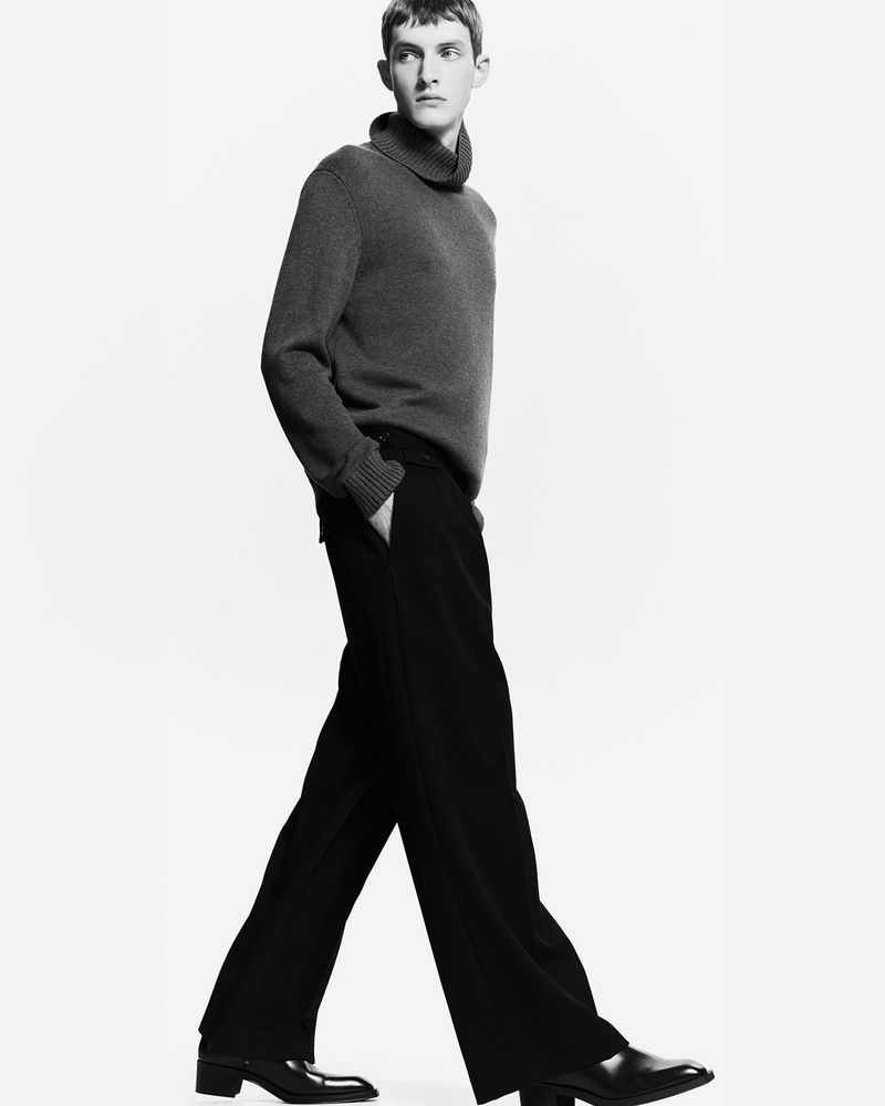 Black and white fashion shot of a slender male model in a turtleneck sweater and black pants, walking against a clean white backdrop.