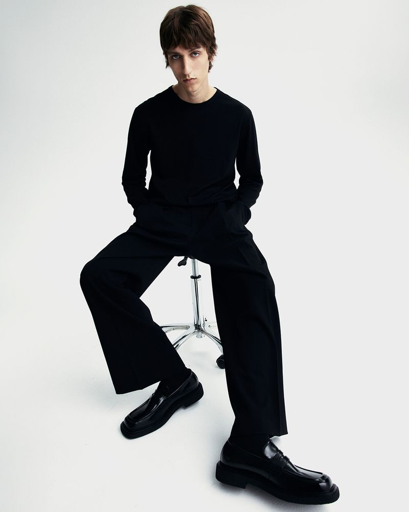A young adult man sits on a stool in a photo studio, wearing a long-sleeve black shirt, black pants, and black loafers against a white backdrop.