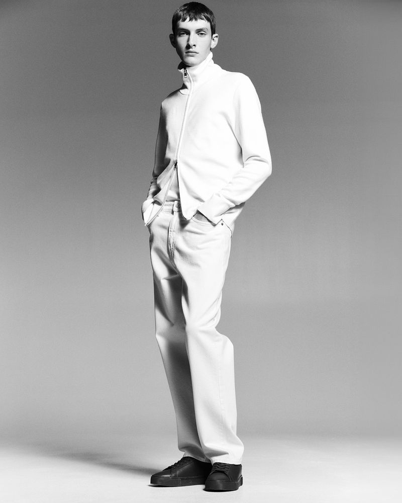 Monochrome studio shot of a young adult male model in an all-white casual outfit, with a jacket, pants and black shoes, hands in pockets.