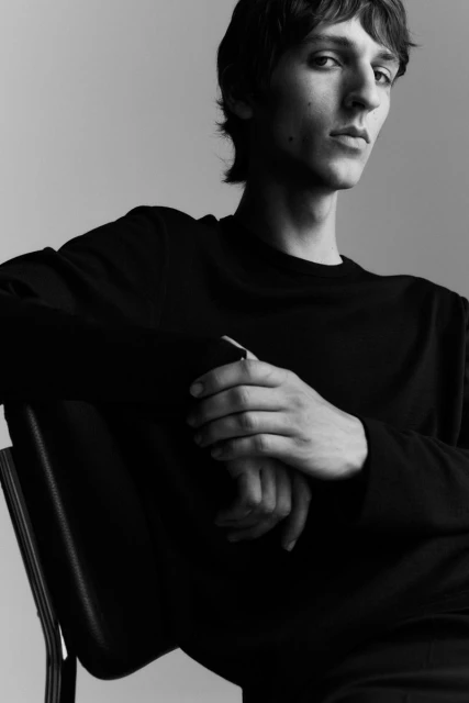 Black and white portrait of an adult male in a dark long sleeve shirt sitting on a chair in front of a plain grey background.