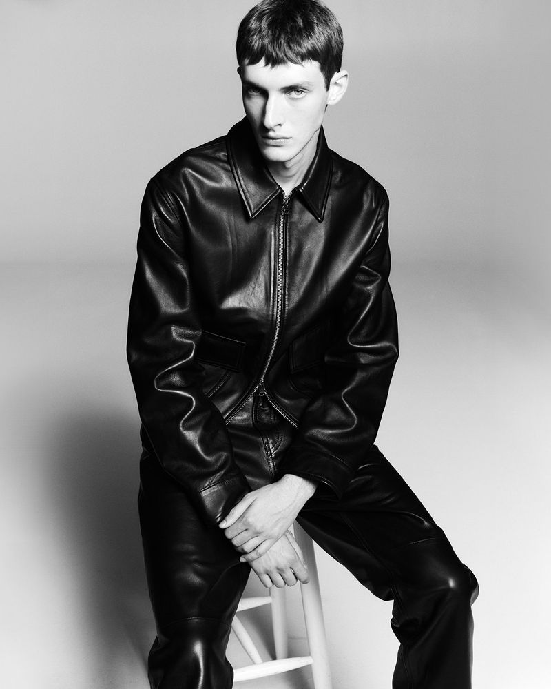 Monochrome portrait of a young male model wearing a black leather jacket seated against a plain neutral background.