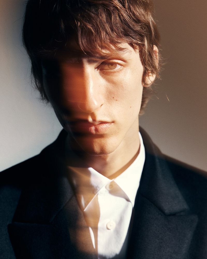 Portrait of a young adult male with brown hair wearing a black coat and white shirt, standing indoors with soft studio lighting.