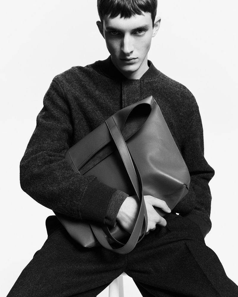 Monochrome fashion portrait of a young adult male model holding a large leather tote bag against a bright white background.
