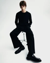Young male model seated on a stool wearing a black long-sleeve top and black trousers against a white seamless background