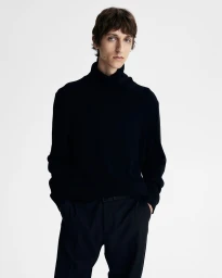 A male model wearing a dark turtleneck sweater and trousers against a plain white studio background.