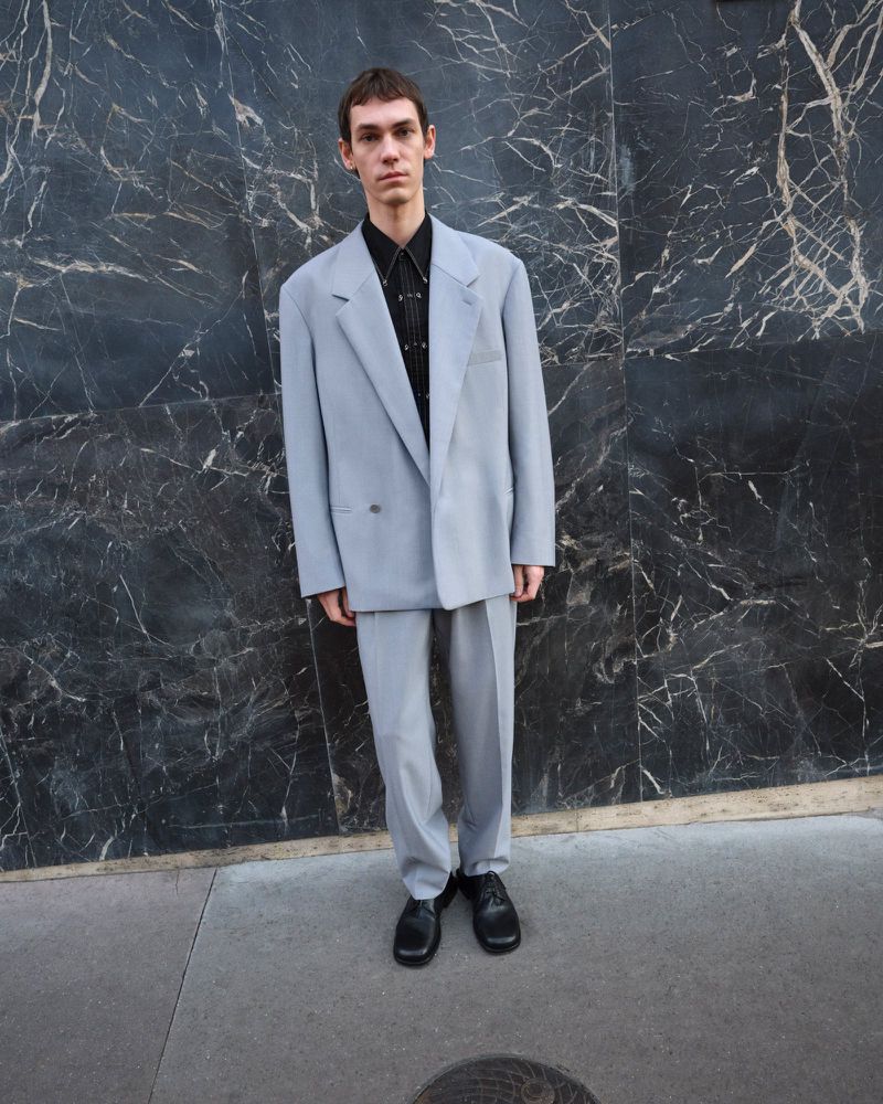 A young adult man poses in an oversized grey suit and black dress shoes against a black marble wall, showcasing a sophisticated style.