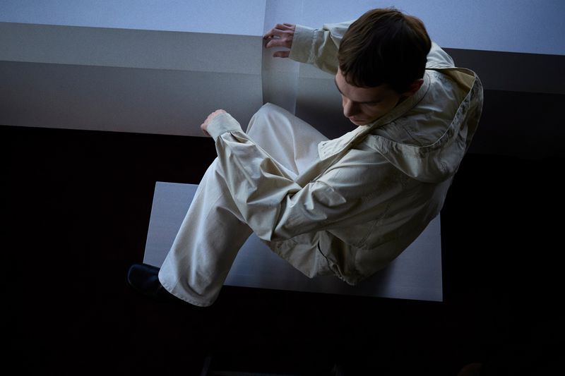 Man in hooded jacket and beige pants sits in a dark corner. Overhead shot shows an eye-level perspective in a minimal environment.