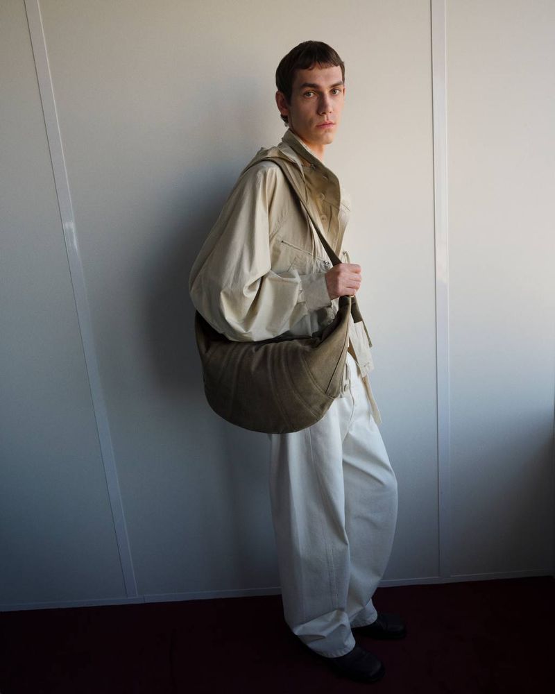 Model wearing a light utility shirt and white trousers carrying a large curved canvas shoulder bag against a neutral studio wall.