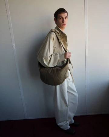 Model wearing a light utility shirt and white trousers carrying a large curved canvas shoulder bag against a neutral studio wall.
