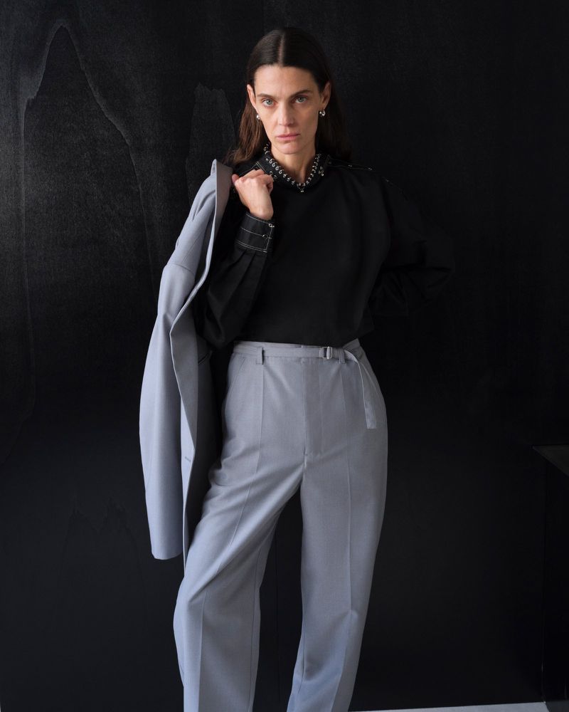 Elegant adult woman in a black studded blouse and light grey suit, standing in front of a dark background in a studio.