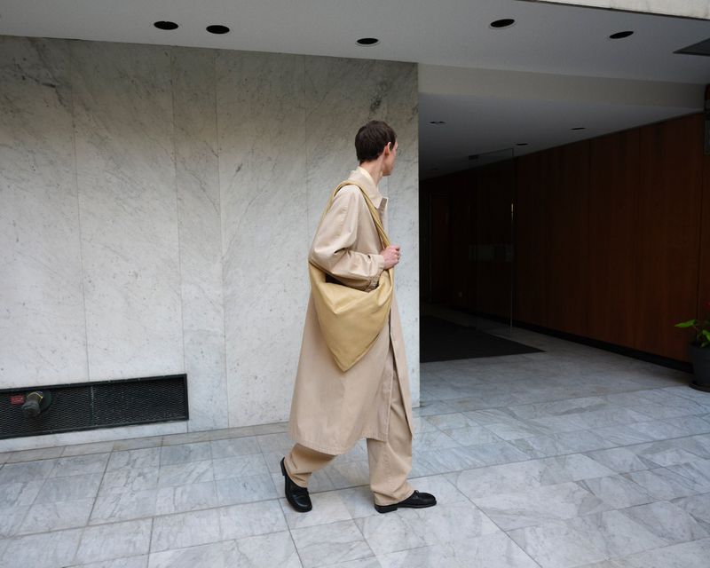 A slender adult walks on a tile floor in a tan overcoat and matching pants, carrying a bag with light reflecting off the material.