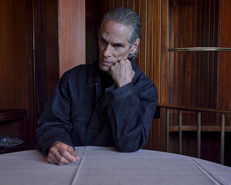 Portrait of a grey-haired adult male wearing a denim jacket, sitting at a table indoors with wood paneling in the background.