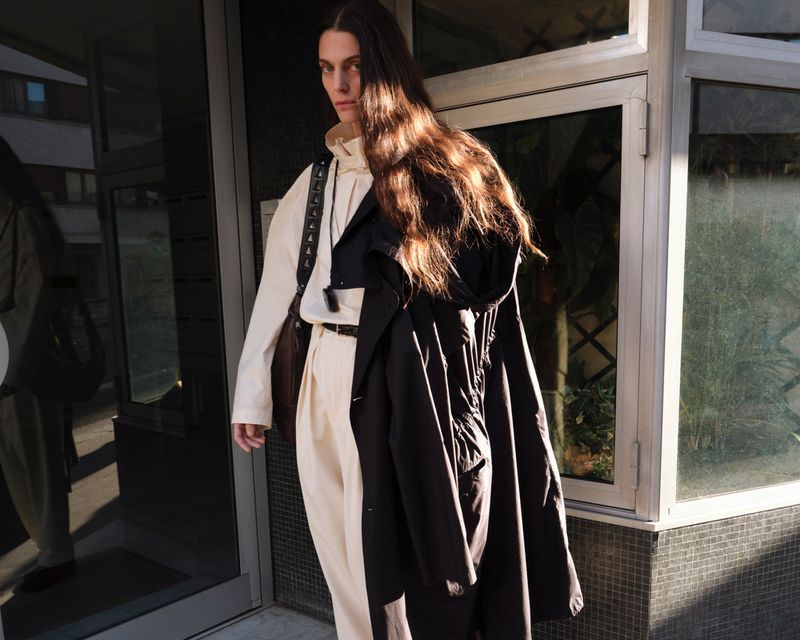 Model standing outdoors wearing an off-white outfit with a dark coat draped over one shoulder and a shoulder bag.