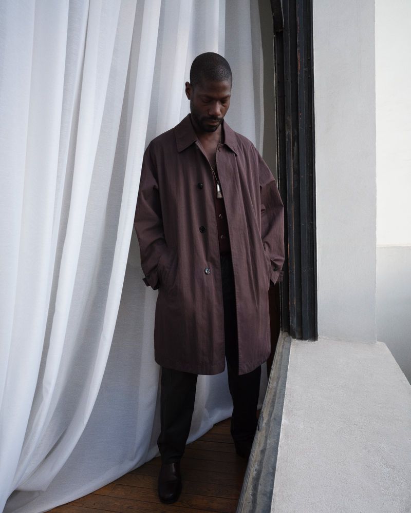 Model in a long brown coat standing next to white curtains near a doorway for Lemaire lookbook