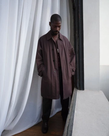 Model in a long brown coat standing next to white curtains near a doorway for Lemaire lookbook