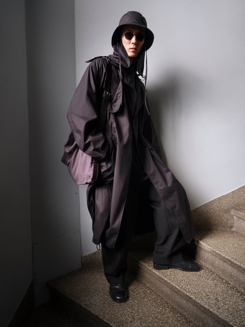 Person in a dark, hooded overcoat with a messenger bag, posing on grey stairs and wall backdrop, casting a shadow.