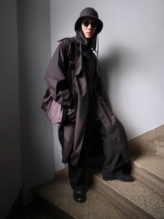 Person in a dark, hooded overcoat with a messenger bag, posing on grey stairs and wall backdrop, casting a shadow.