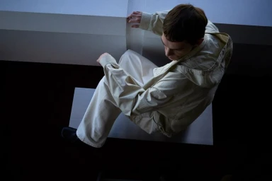 High-angle shot of a male model wearing cream-colored hooded jacket and wide trousers seated on a white platform